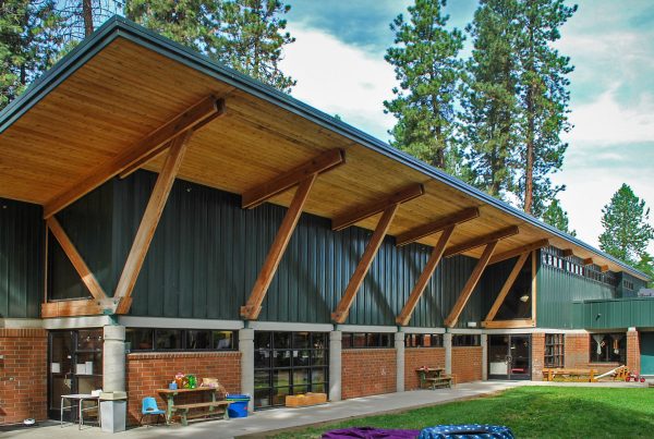 Exterior courtyard view of the North Idaho College Children's Center in Coeur d'Alene, Idaho, showing a modern building with exposed timber supports, green metal siding, red brick walls, and outdoor play areas beneath a canopy of pine trees.