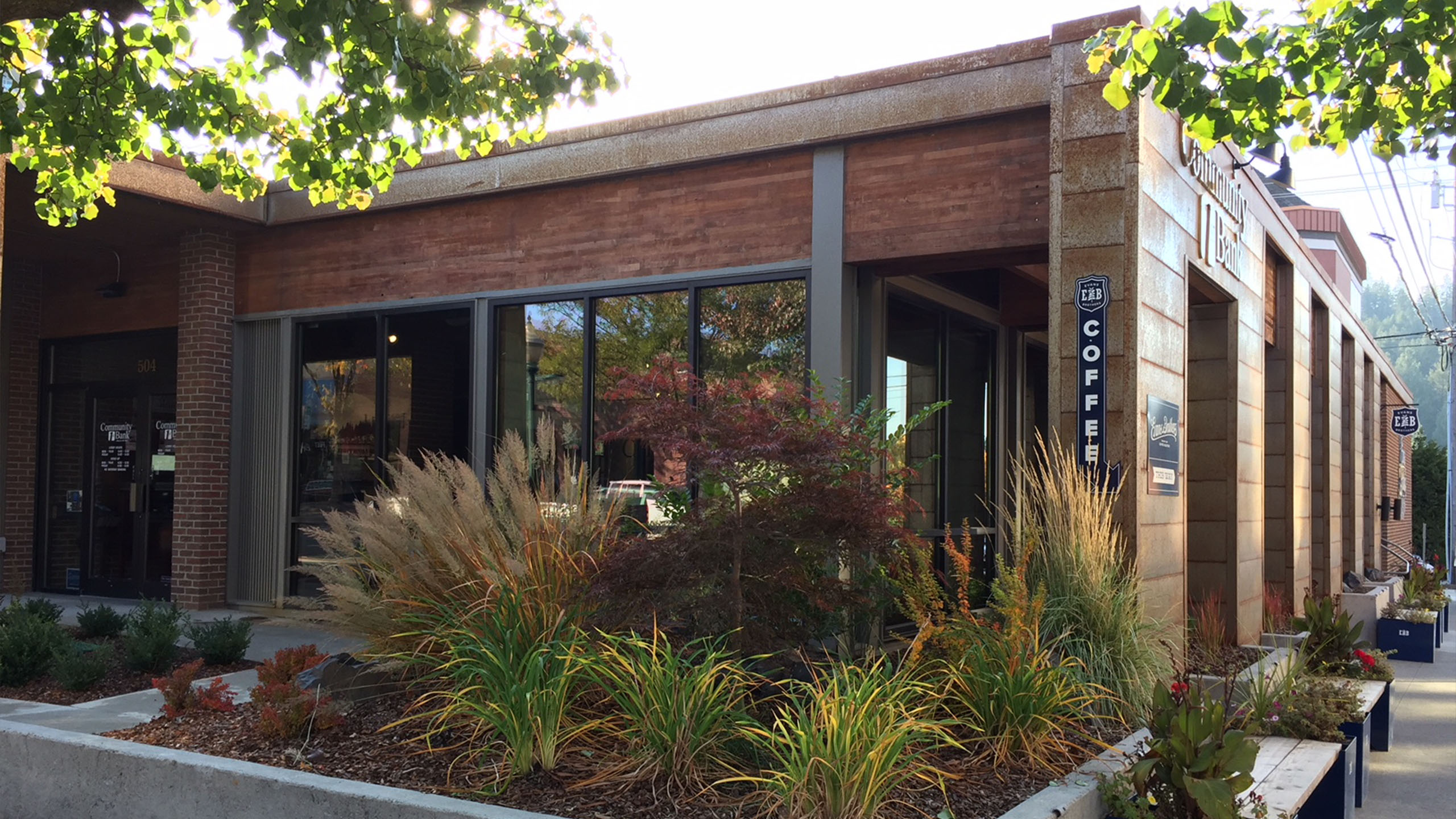 Corner view of Community 1st Bank in downtown Coeur d’Alene, featuring weathered steel facade, landscaped planters, and street-facing coffee shop.