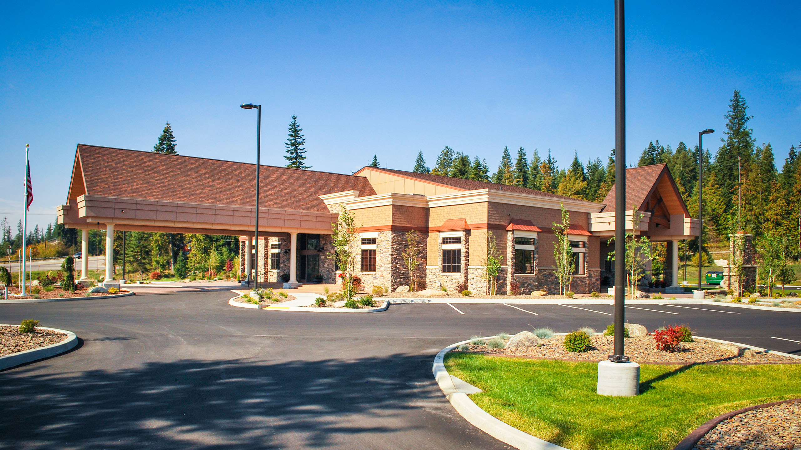 Front view of Priest River Event Center in Idaho, showcasing its lodge-inspired architecture with stone and wood accents, gabled rooflines, and a large covered drop-off area, surrounded by forest and landscaped parking.