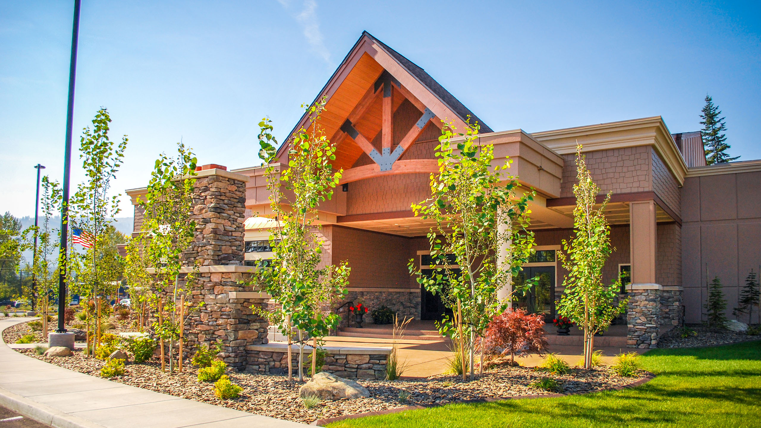 Close-up view of the exterior patio of Priest River Event Center in Idaho, featuring a gabled roof with exposed timber trusses, stone columns, and landscaped surroundings with young trees and flowering plants.