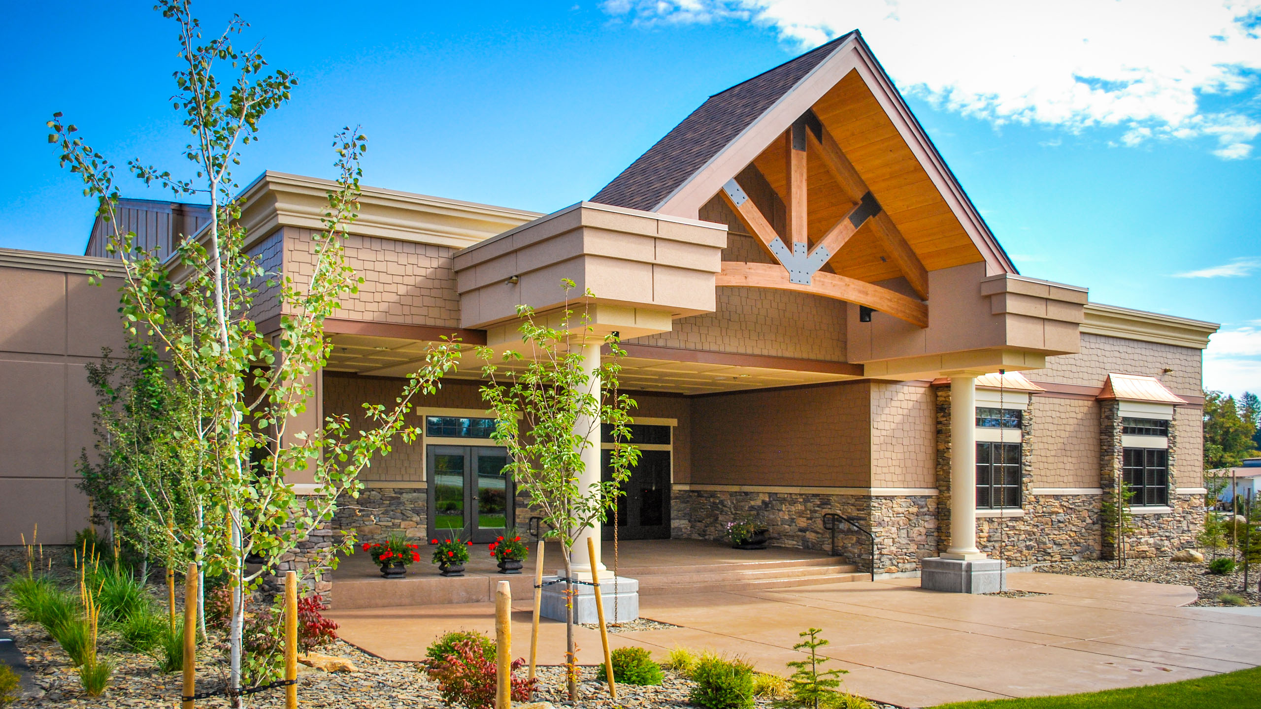 Exterior view of Priest River Event Center in Idaho, showing an exterior patio space with a gabled roof supported by columns, exposed timber trusses, stone and shingle siding, and surrounding landscaped plantings.