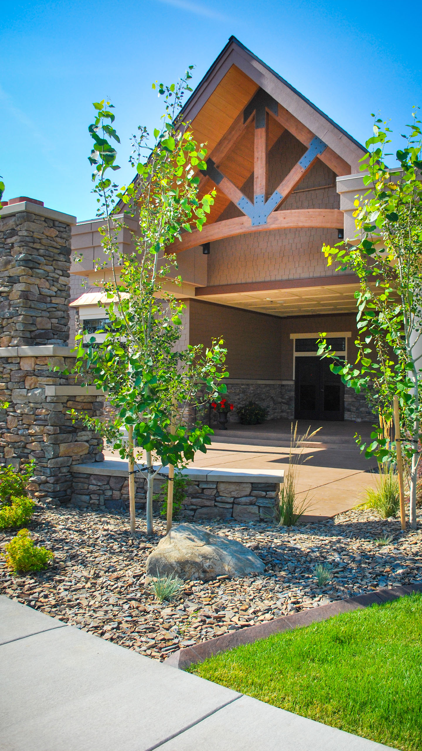 Angled view of an entrance at Priest River Event Center in Idaho, featuring exposed timber trusses, stone columns, and young landscaping under a clear blue sky.