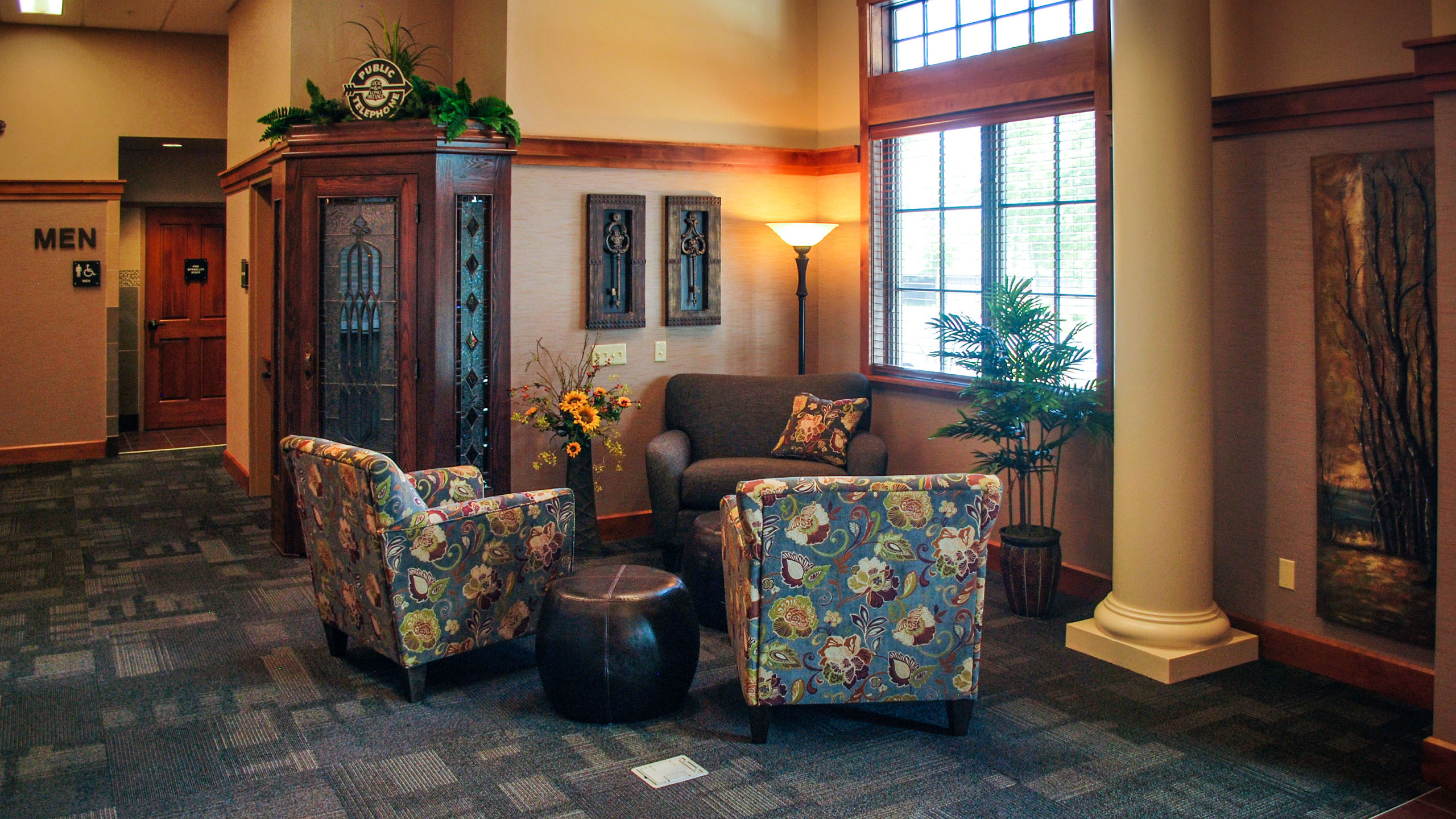 Cozy seating nook at the Priest River Event Center in Idaho, featuring floral-patterned armchairs, a small sofa, decorative floor lamp, and a vintage phone booth with stained glass panels, near large windows and restrooms.