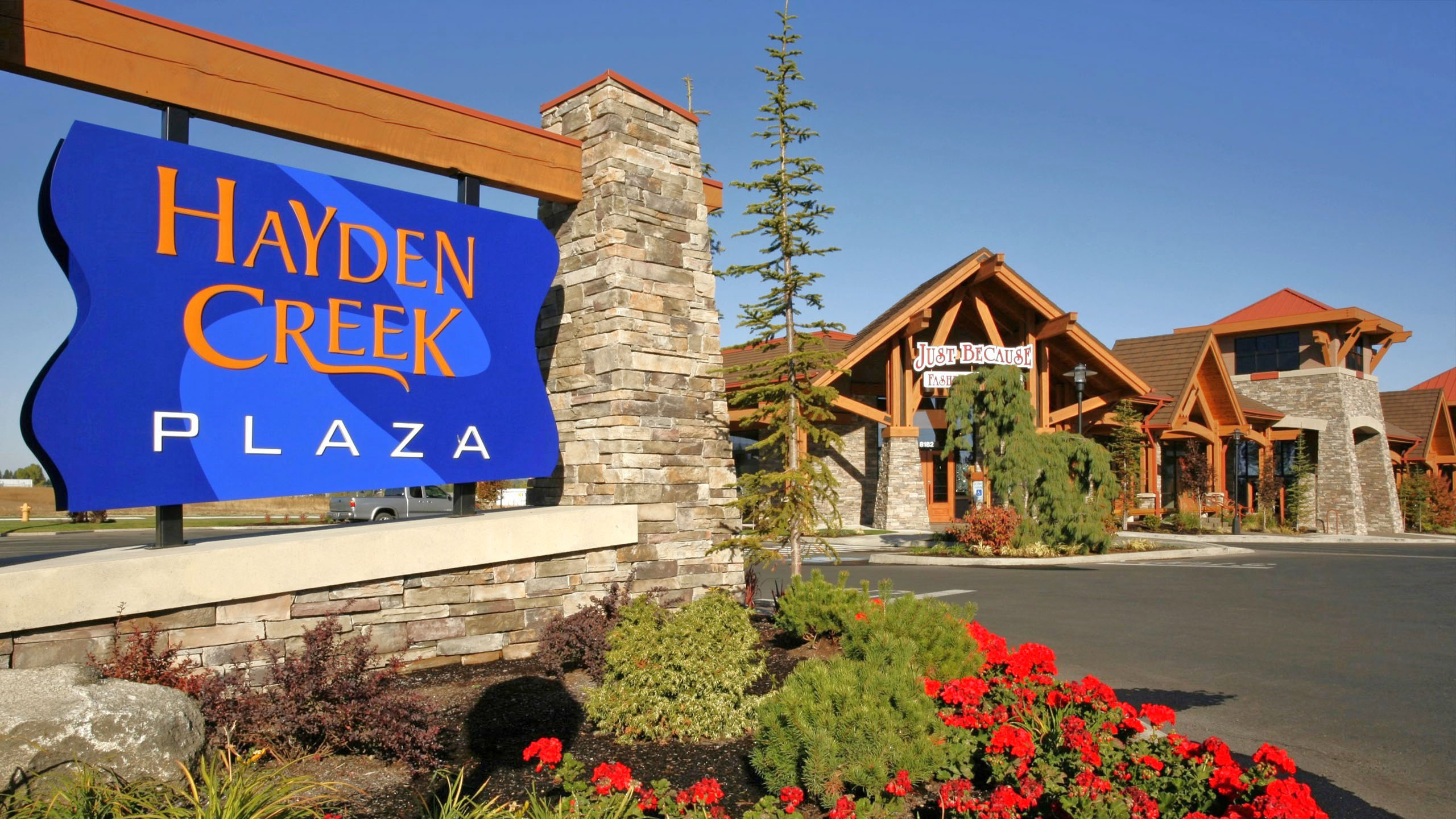 Hayden Creek Plaza entrance sign with landscaped flowers, stone pillar, and timber-framed retail buildings in the background in Hayden, Idaho.
