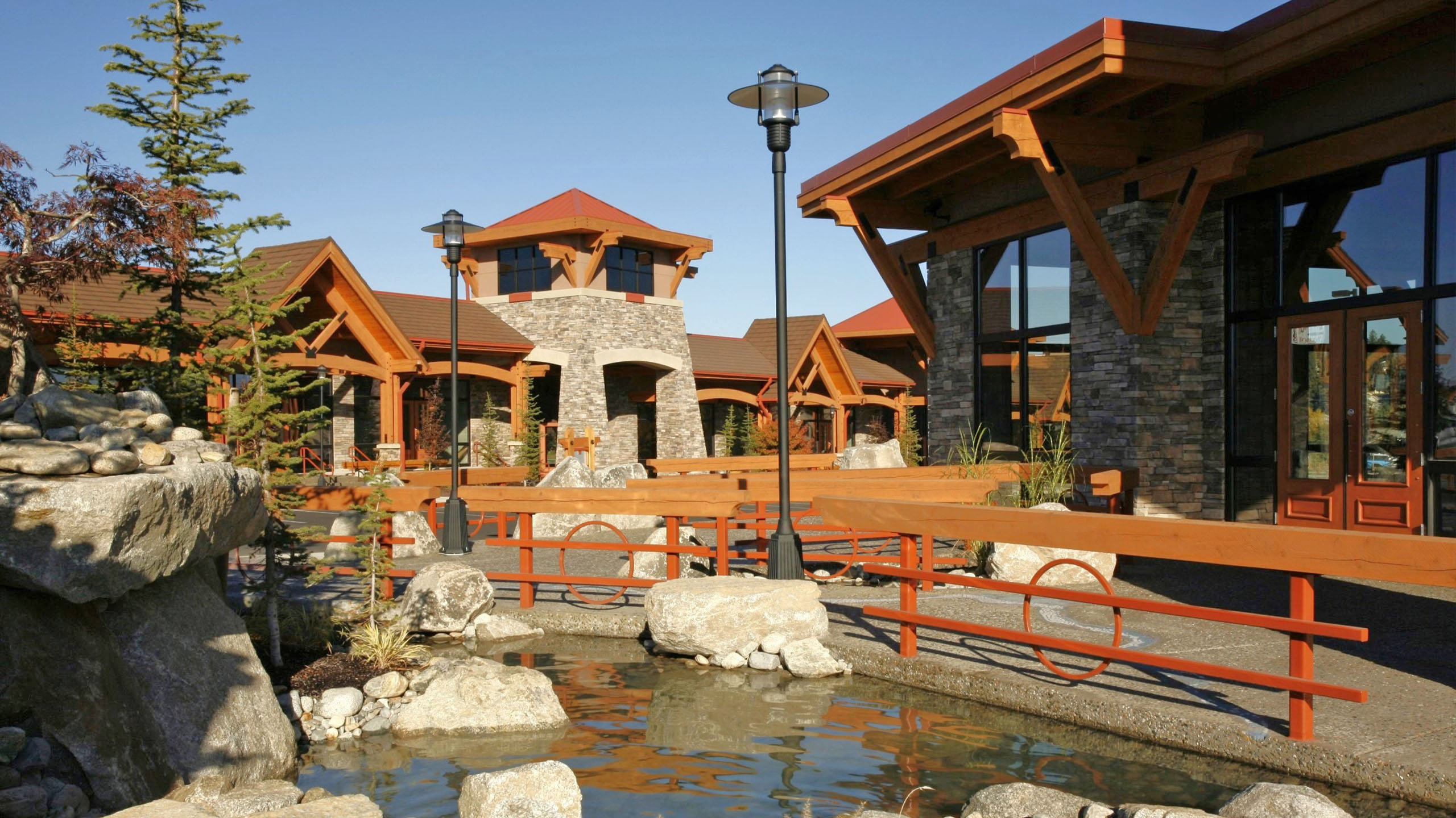 Stone and timber retail buildings at Hayden Creek Plaza in Hayden, Idaho, with a landscaped water feature and pedestrian bridge in the foreground.