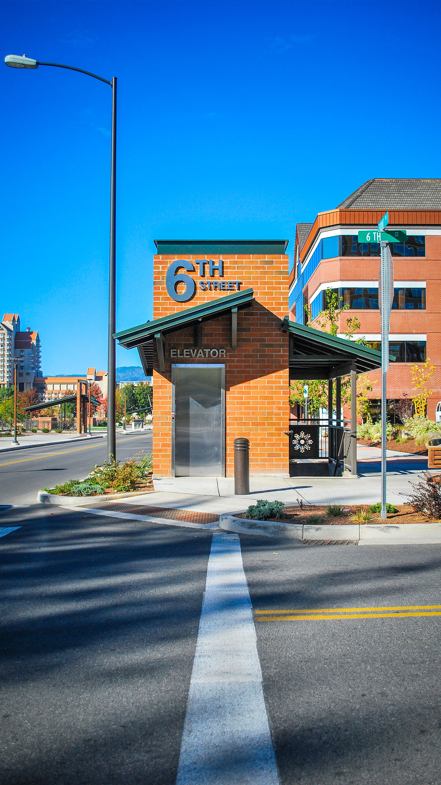 A red brick elevator structure marked "6th Street" with a green metal roof, providing pedestrian access between street level and McEuen Park in Coeur d’Alene, Idaho. The surrounding area includes modern buildings, landscaping, and clear signage under a vibrant blue sky.