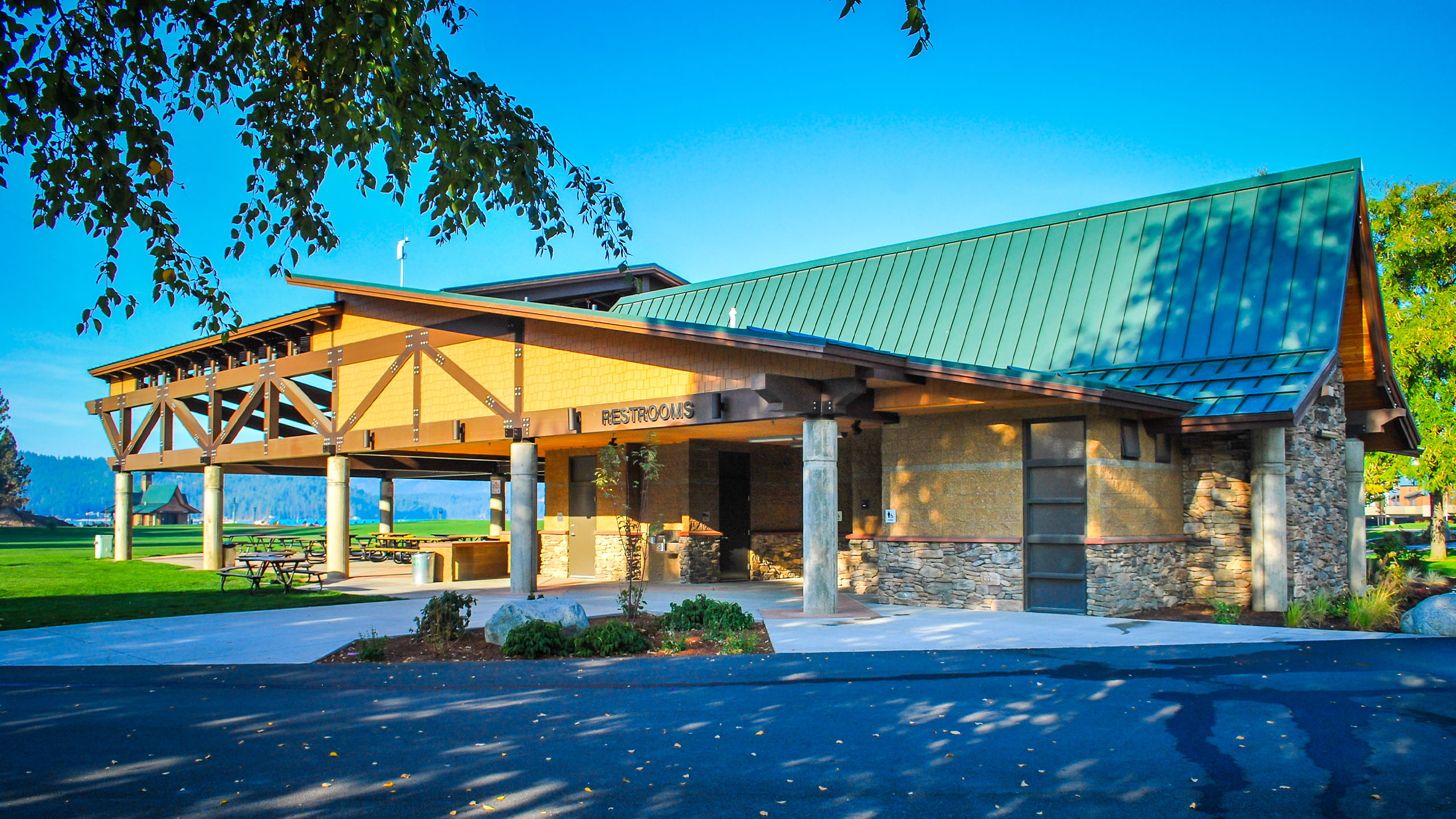 A pavilion-style public restroom facility at McEuen Park in Coeur d’Alene, Idaho, featuring a green metal gabled roof, stone and wood exterior, and open-air picnic area under extended beams. The building is surrounded by grassy parkland with lake and mountain views in the distance.