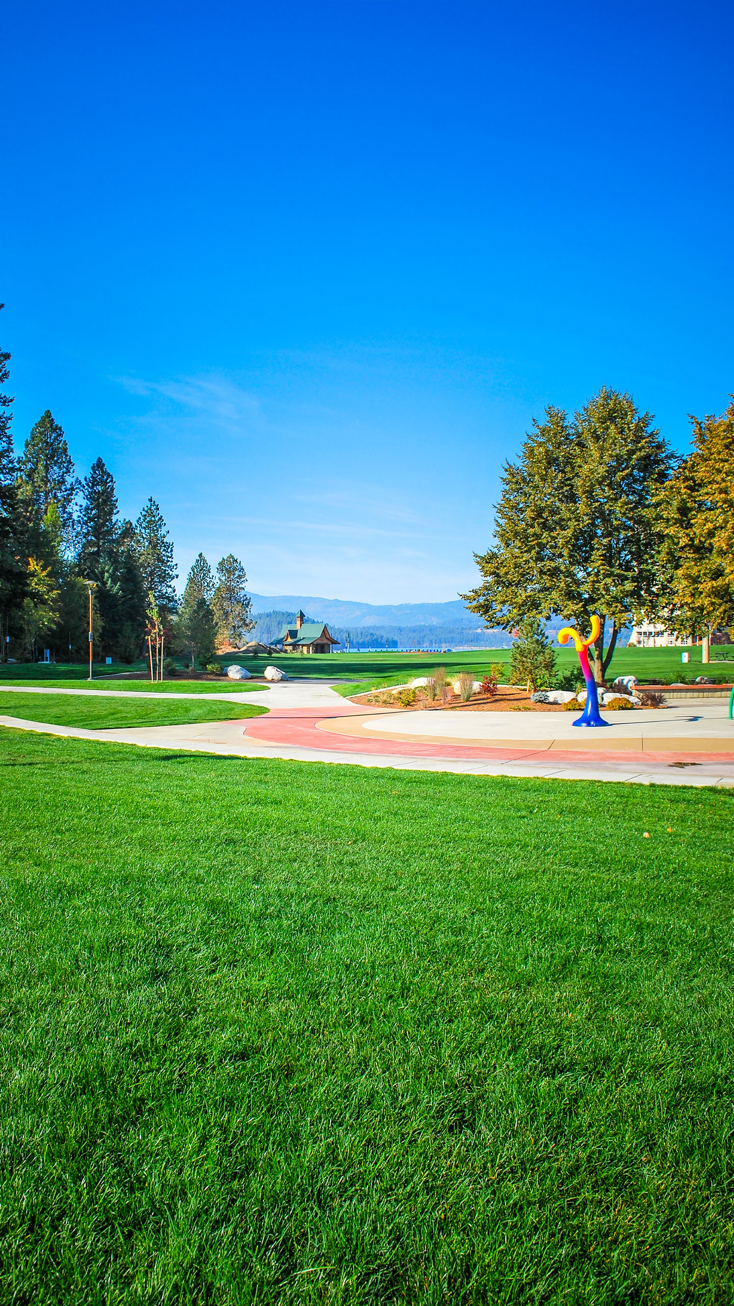 A wide expanse of lush green lawn at McEuen Park in Coeur d’Alene, Idaho, with a colorful splash pad feature to the right and a tree-lined walkway leading toward a lakeside pavilion in the distance. Forested hills and blue skies form the scenic backdrop.