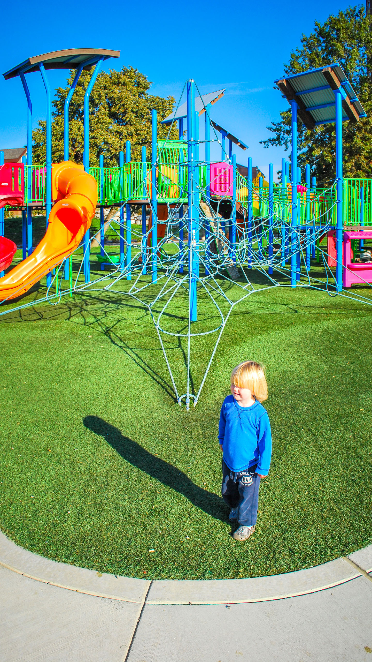 A young child stands near a colorful playground at McEuen Park in Coeur d’Alene, Idaho. The playground features vibrant blue, green, pink, and orange climbing structures, slides, and rope nets, set on artificial turf under a clear blue sky.