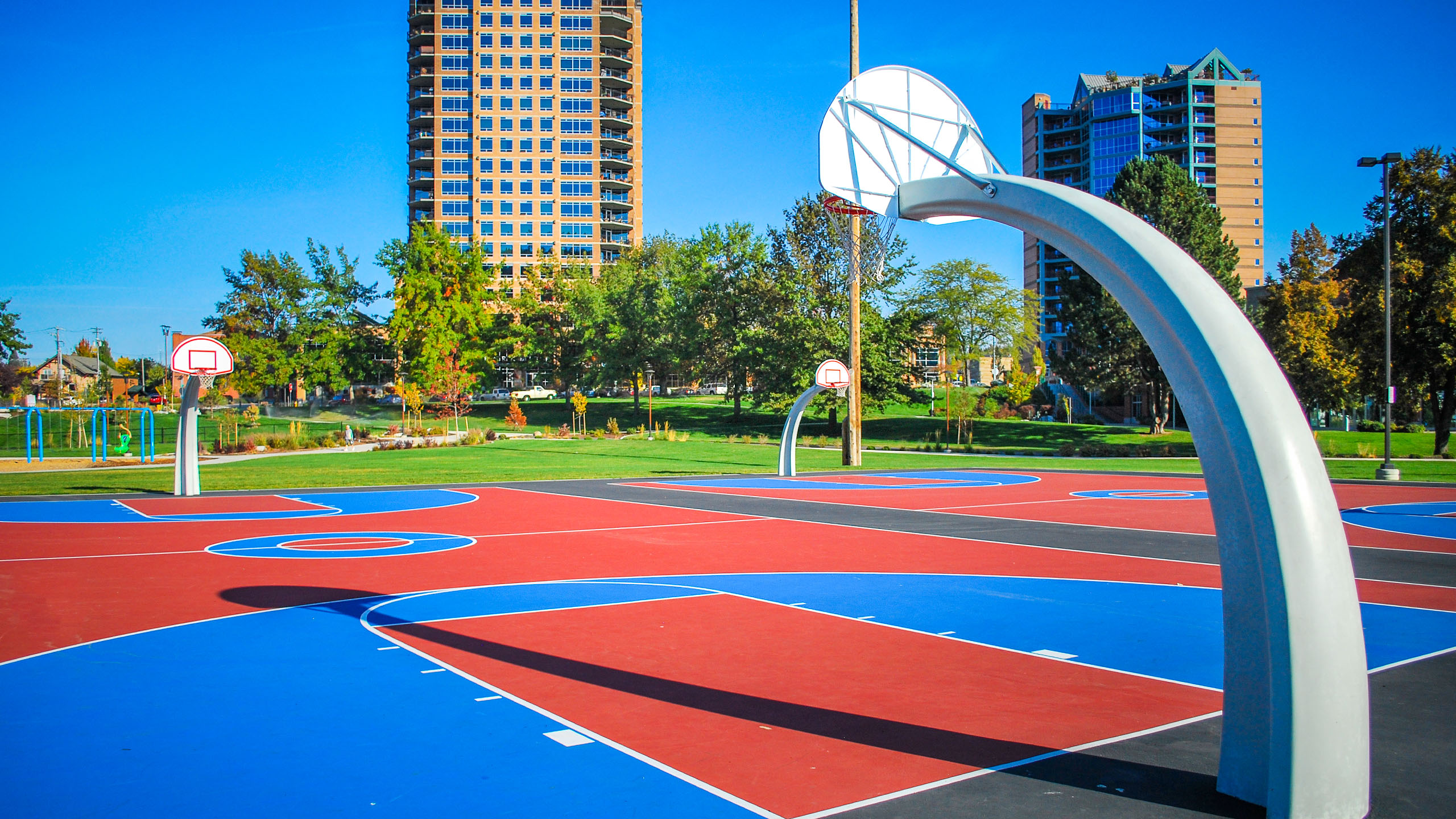 View of the colorful outdoor basketball courts at McEuen Park in Coeur d’Alene, Idaho, featuring bold red, blue, and black court markings with modern curved white basketball hoops. In the background, tall residential buildings rise behind a lush line of trees under a vibrant blue sky.