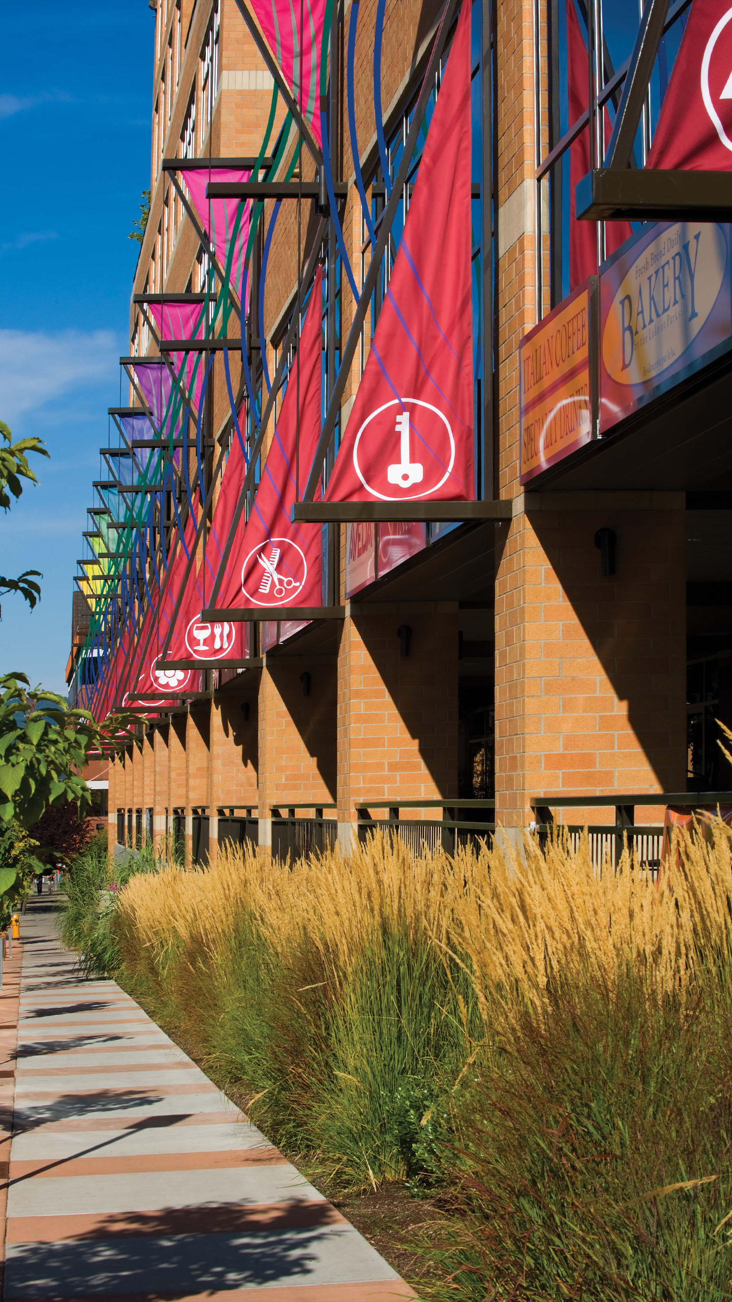 Sidewalk-level view of the Parkside building in Coeur d’Alene, Idaho, showing tall ornamental grasses, colorful vertical banners with icons, and brick columns supporting the upper structure. Small businesses and signage are visible at the street level.