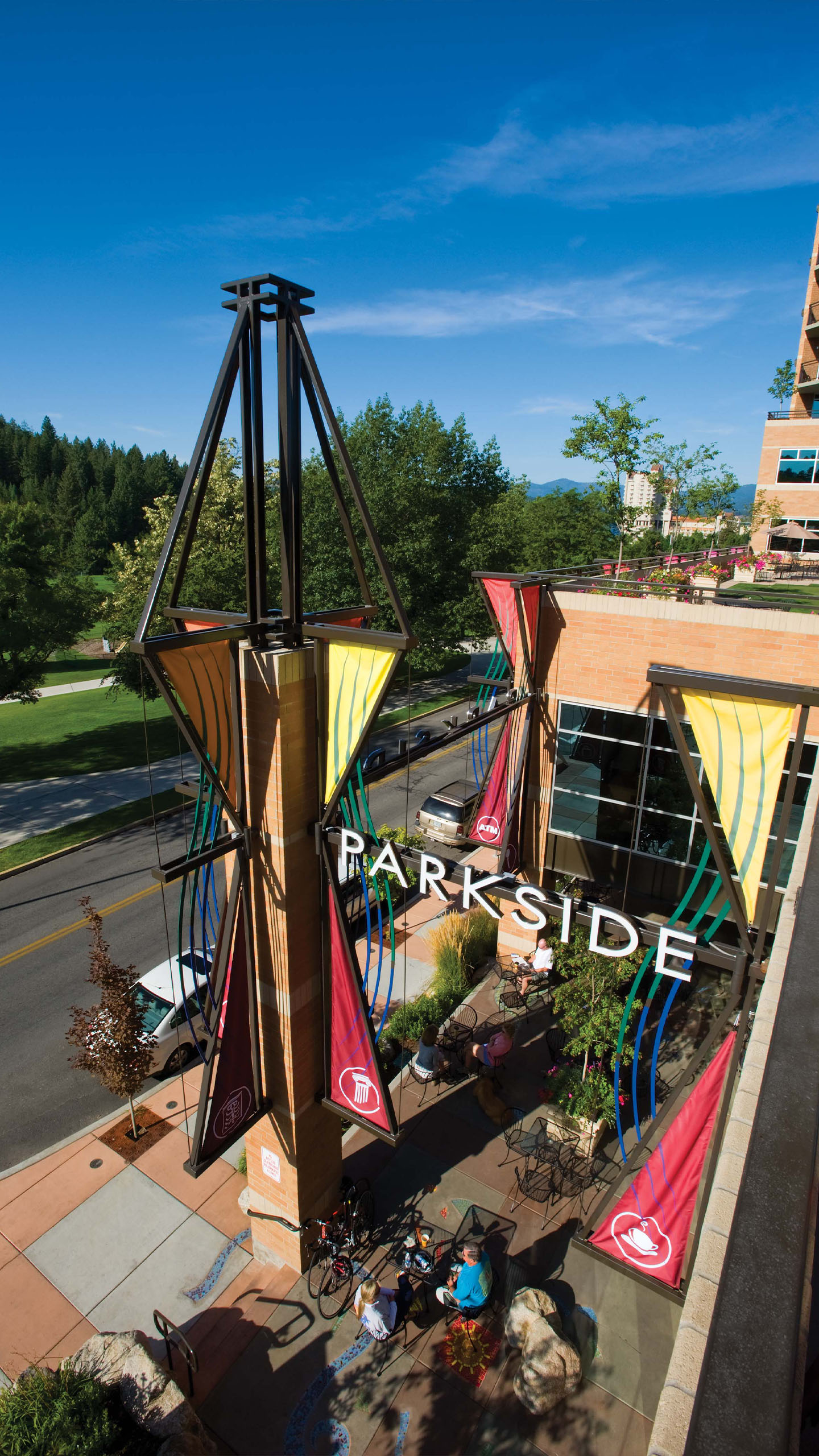 Daytime view of the Parkside building in Coeur d’Alene, Idaho, showing colorful sculptural columns with red, yellow, and blue accents framing the “PARKSIDE” sign. People are gathered at outdoor tables below, with trees, a park, and blue skies in the background.