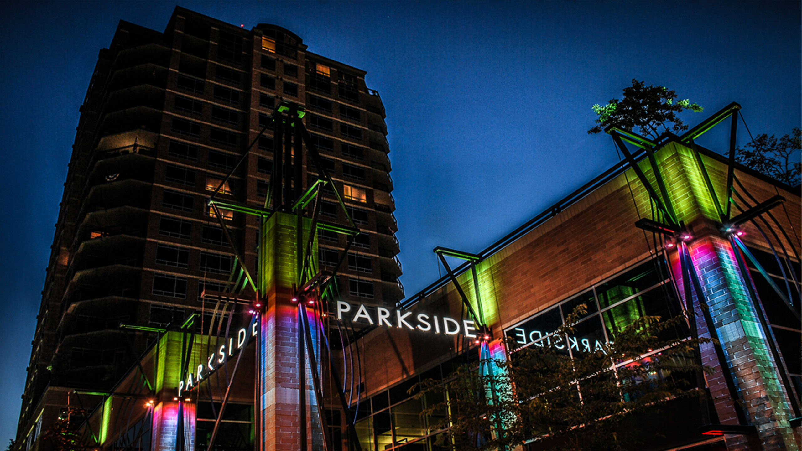 Night view of the Parkside building in Coeur d’Alene, Idaho, featuring dramatic architectural lighting in green, blue, and magenta illuminating structural columns, with the "PARKSIDE" signage glowing along the facade and the tower rising above into the twilight sky.