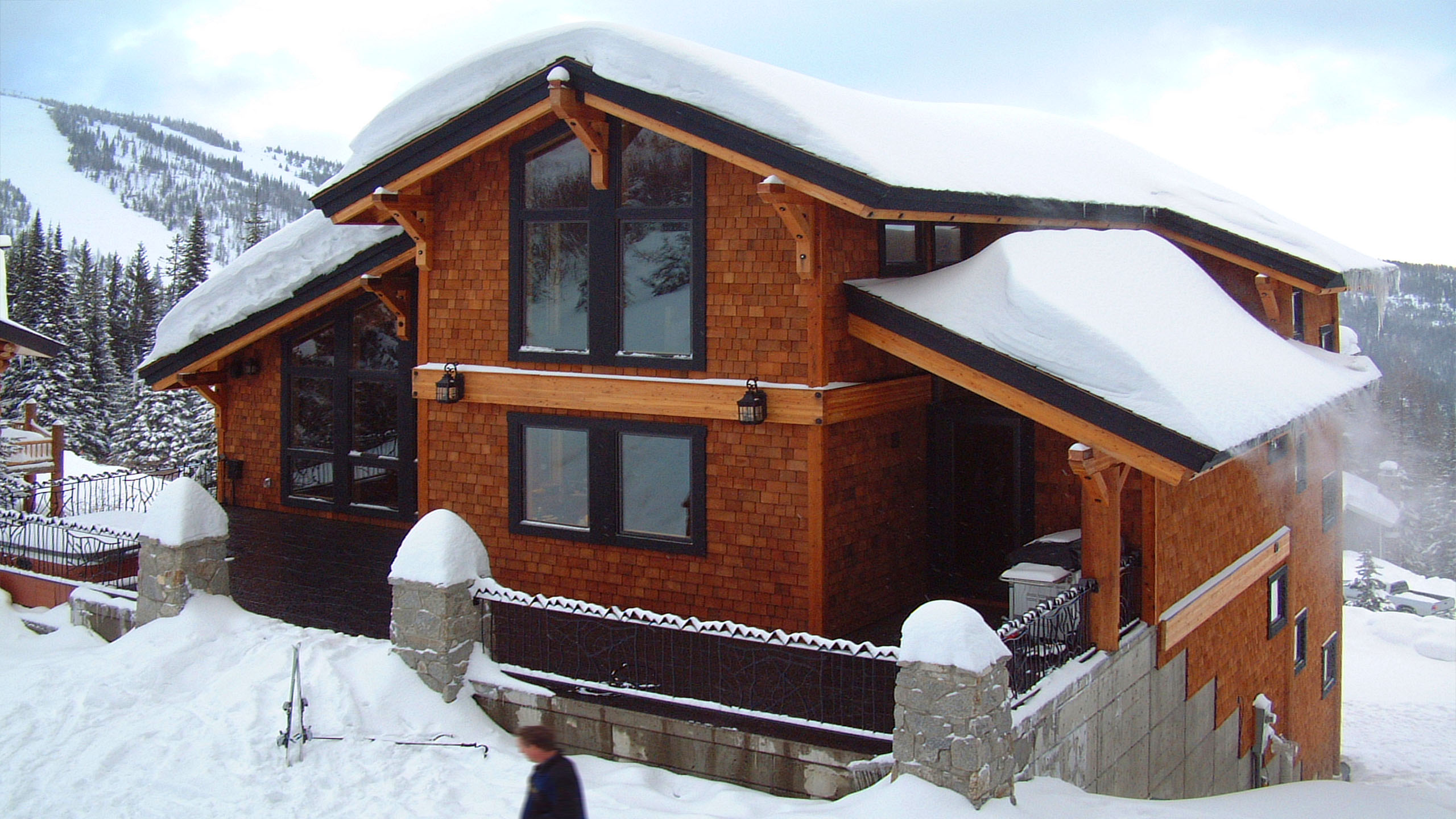 Side view of Schweitzer Ski Cabin in Sandpoint, Idaho, featuring cedar shingle siding, steep snow-covered roofs, and black-framed windows nestled into a snowy mountain slope.