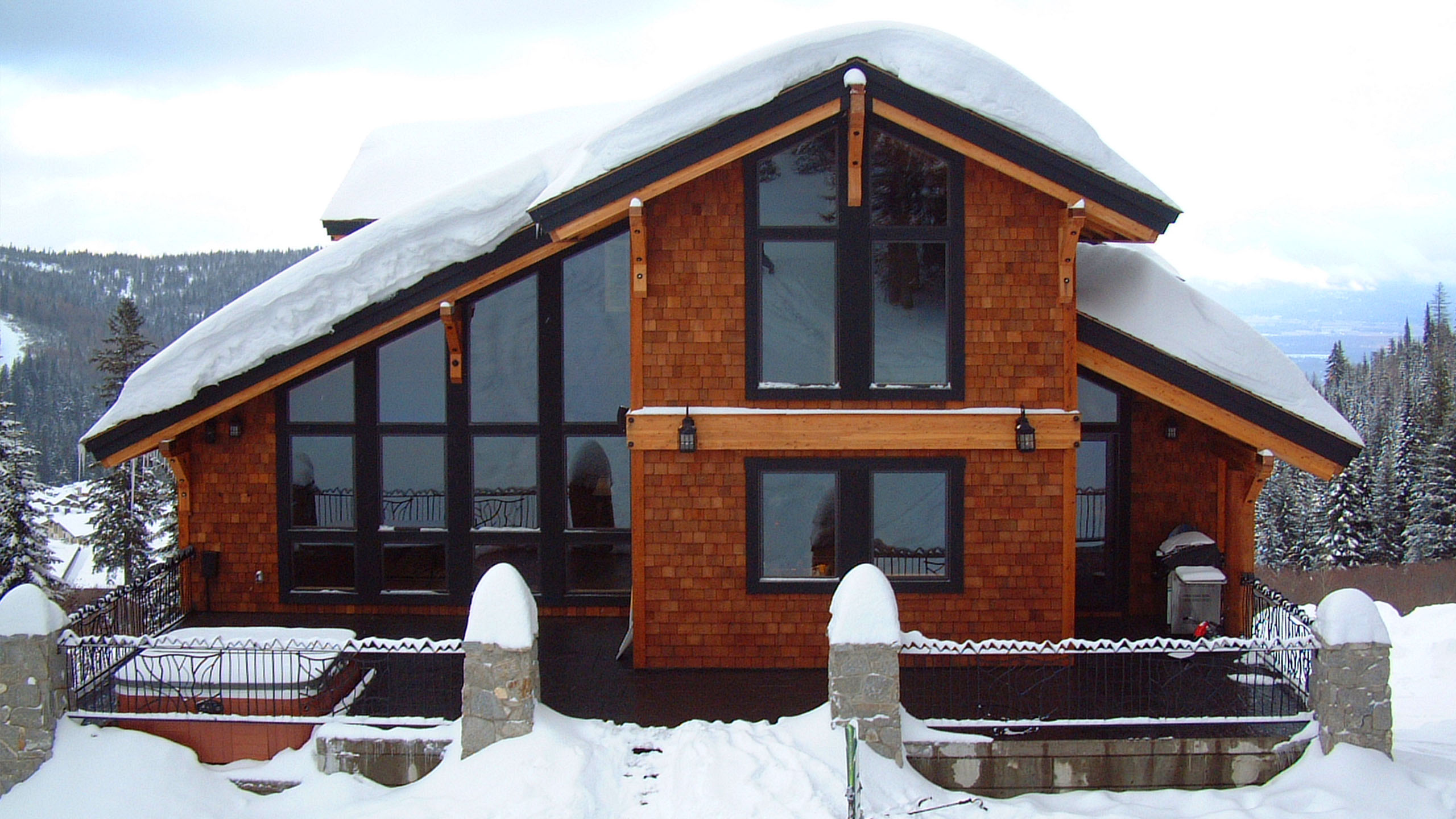 Back elevation of Schweitzer Ski Cabin in Sandpoint, Idaho, with steep snow-covered rooflines, cedar shingles, and large black-trimmed windows overlooking snowy alpine terrain.