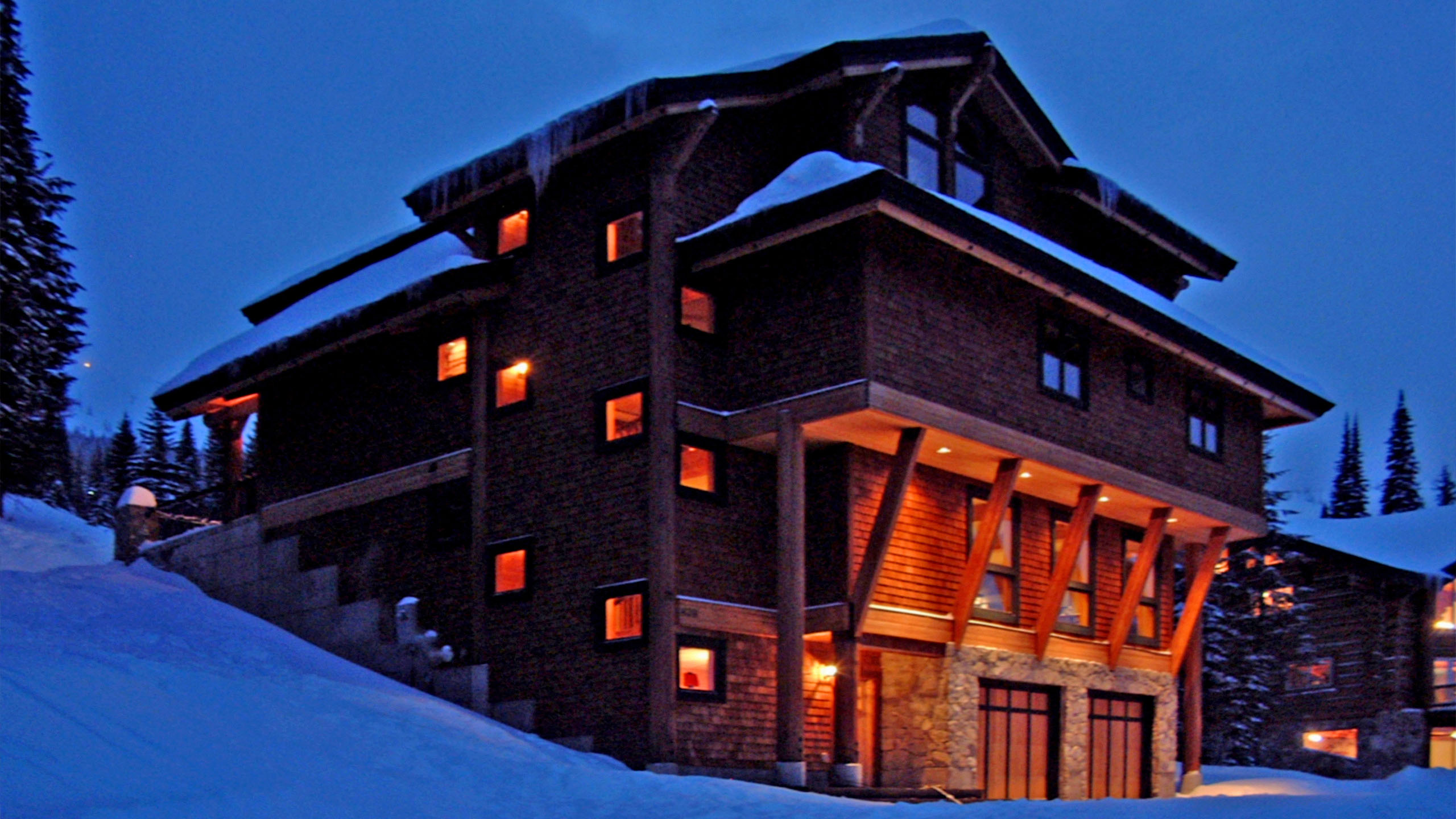 Schweitzer Ski Cabin exterior at dusk in Sandpoint, Idaho, with warm interior lighting glowing through wood-shingled facade and timber-framed entrance, nestled in a snowy mountain landscape.
