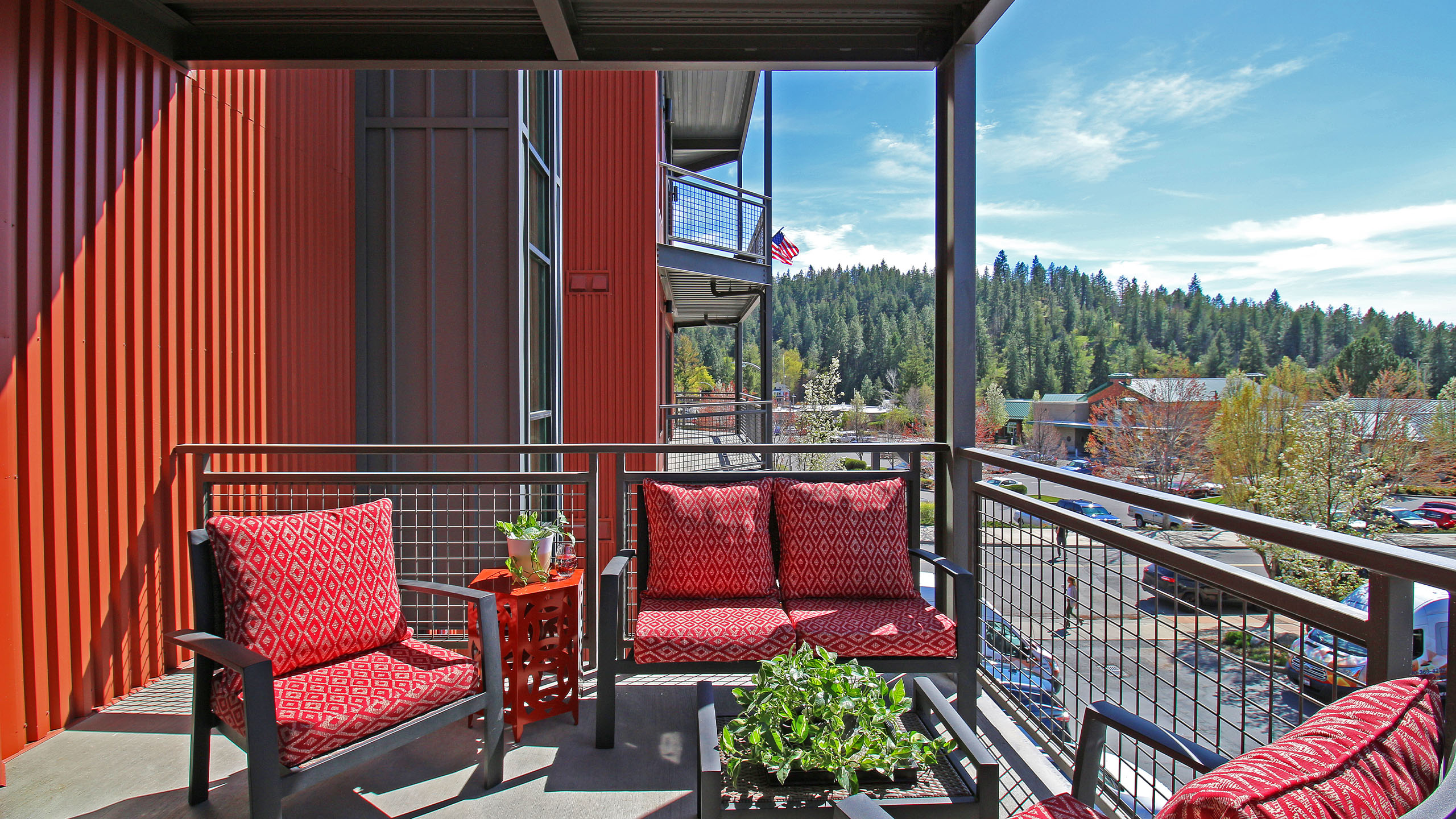 A modern condominium balcony at Seven27 in Coeur d’Alene, Idaho, featuring red metal siding, black-framed furniture with red geometric cushions, and potted plants. The balcony overlooks a parking lot and surrounding buildings, with a view of pine-covered hills and a clear blue sky in the background. An American flag hangs in the distance from another unit.