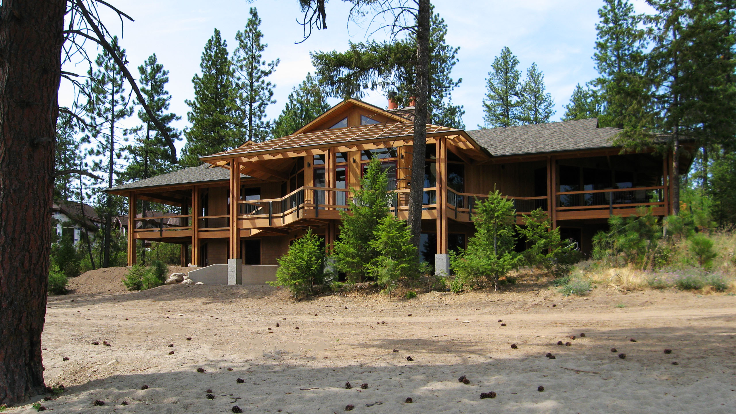 Wide-angle view of Spokane River Residence, a custom timber-framed home with expansive upper-level decking, exposed beams, and large windows, nestled in a wooded landscape along the Spokane River in Post Falls, Idaho.