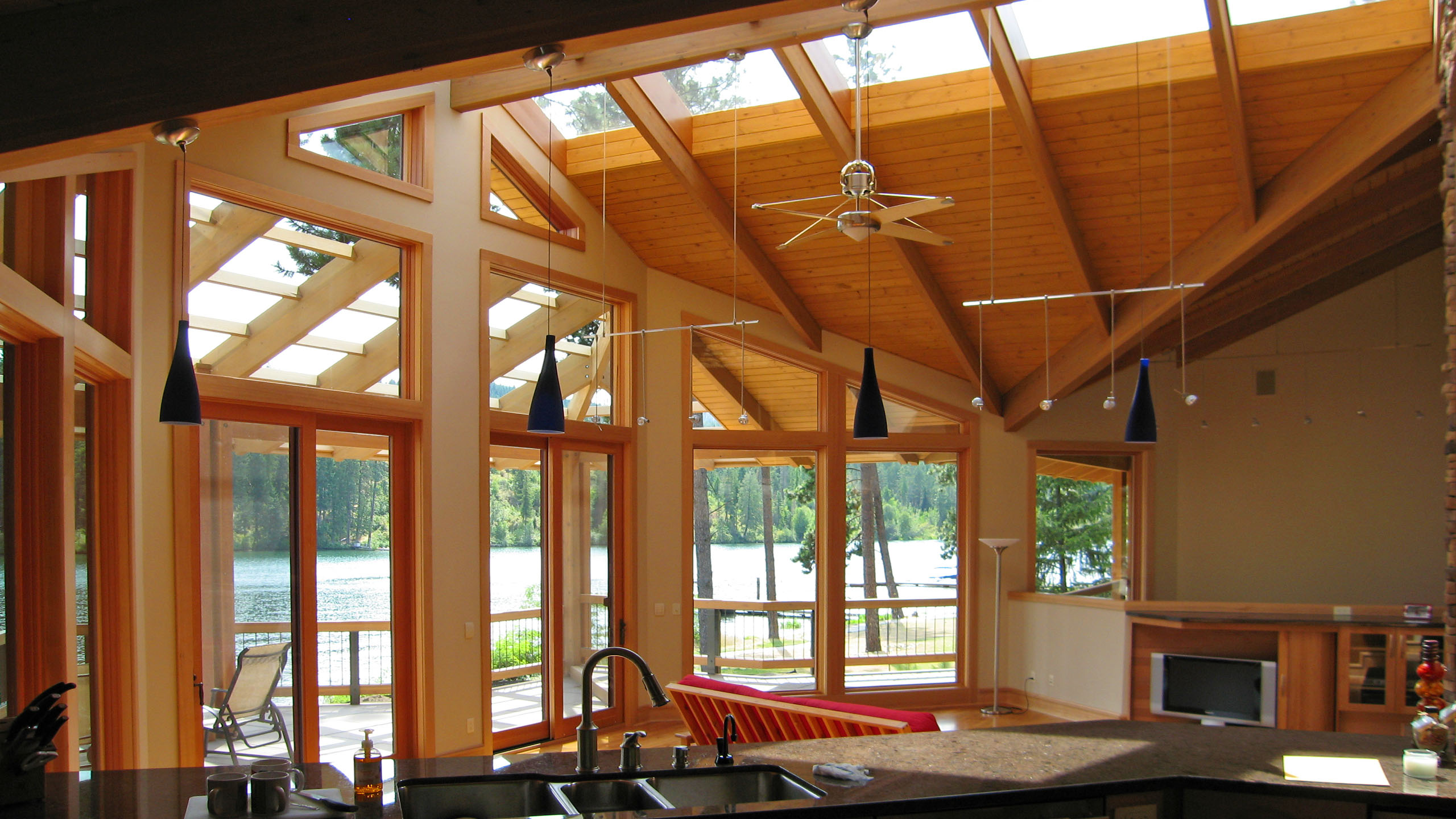 Interior view of Spokane River Residence featuring a vaulted wood ceiling with skylights, large picture windows overlooking the Spokane River, modern pendant lighting, and a kitchen island with a sink in the foreground.