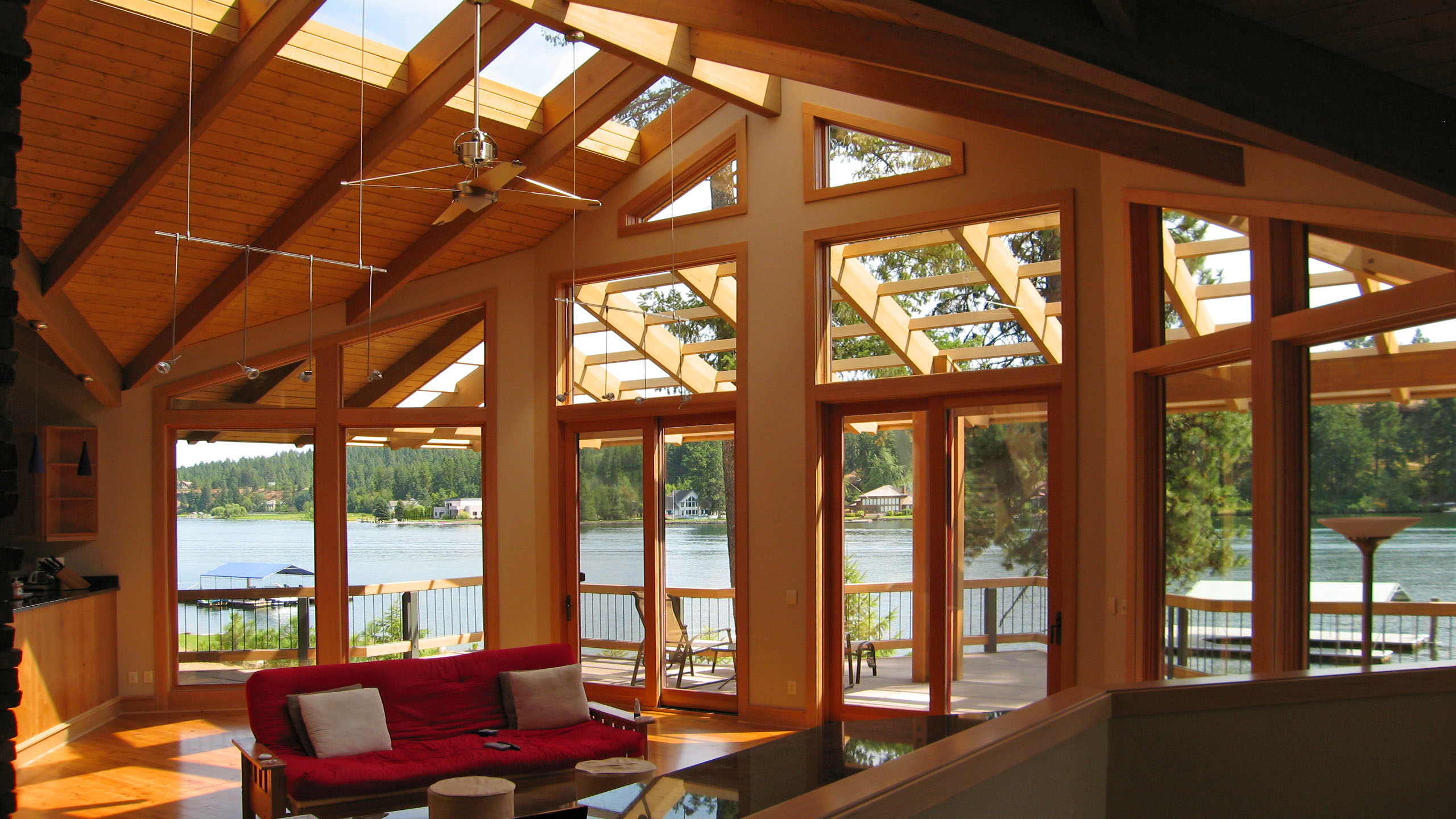 Interior of Spokane River Residence featuring floor-to-ceiling windows and sliding glass doors that open to a deck overlooking the Spokane River, with exposed wood beams, vaulted ceilings, and a red couch in the foreground.