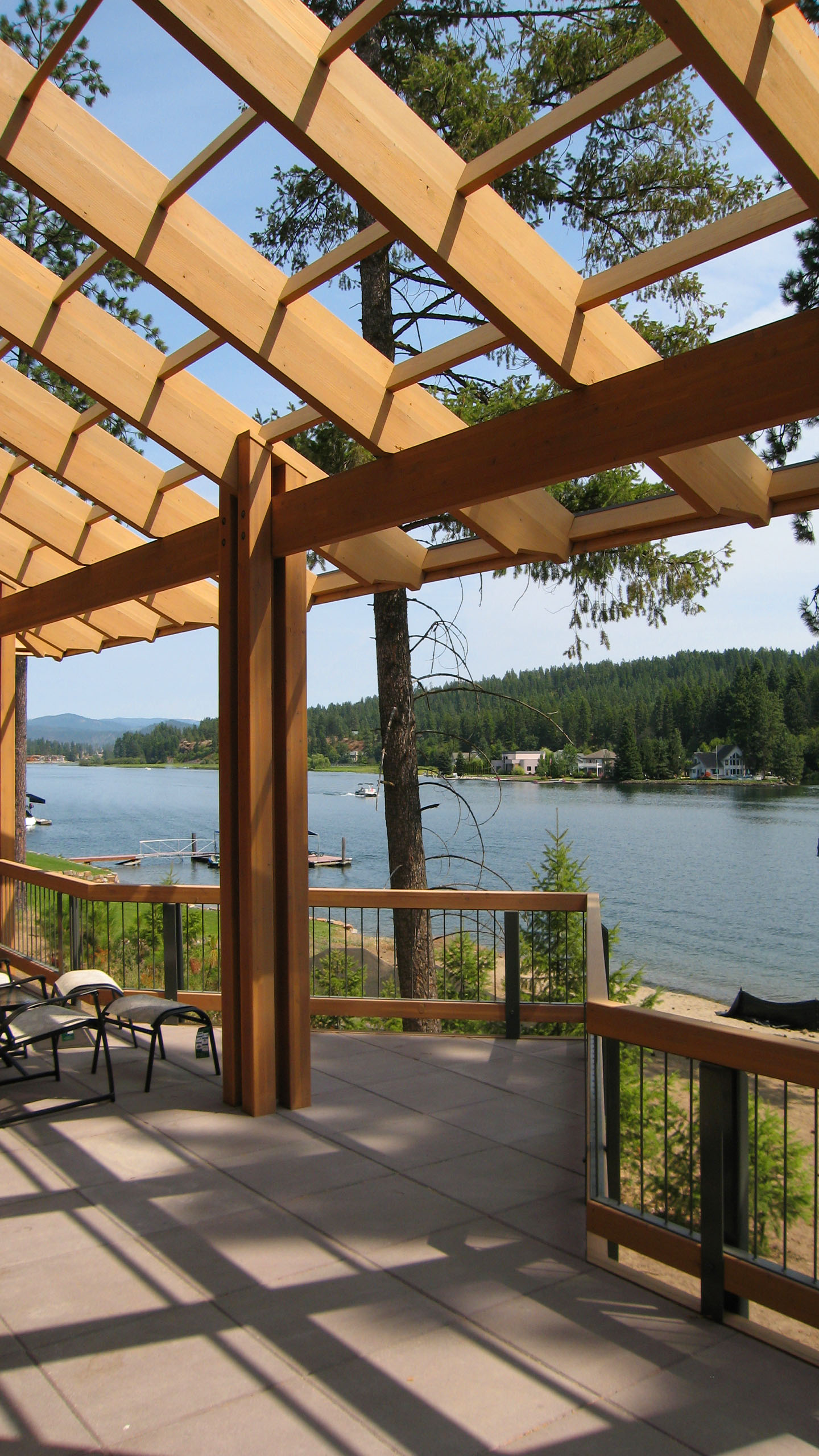 Covered deck at Spokane River Residence overlooking the Spokane River, featuring exposed timber framing and lounge seating with a view of the water and surrounding forested hills.