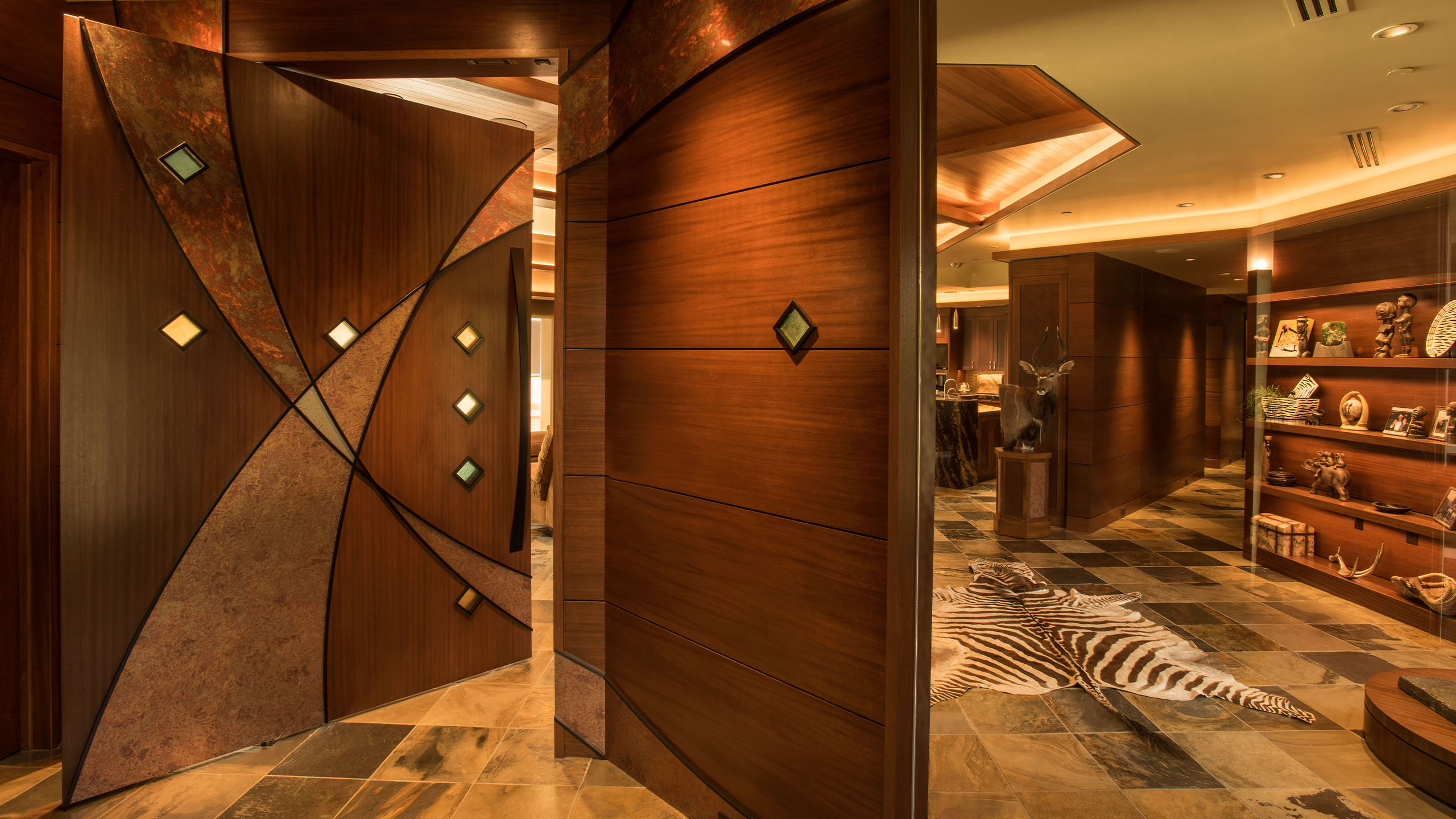 Interior entryway of the Terrace Condo in Coeur d’Alene, Idaho, showcasing a custom curved wood pivot door with copper accents and embedded glass tiles. The space features rich wood finishes, a stone tile floor with a zebra hide rug, built-in display shelving, and ambient lighting that highlights the detailed craftsmanship and luxury throughout.