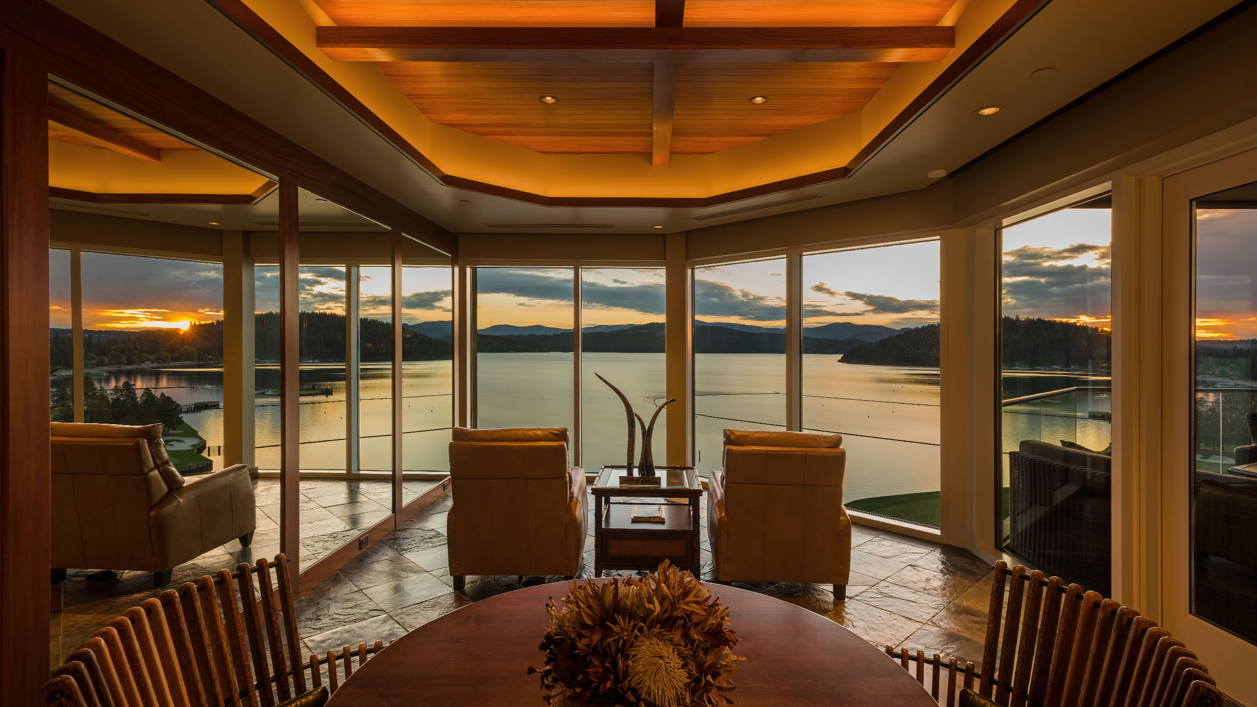 Elegant living space in the Terrace Condo in Coeur d’Alene, Idaho, featuring floor-to-ceiling windows with panoramic views of Lake Coeur d’Alene at sunset. The room includes leather lounge chairs, a round wood dining table, and warm ambient lighting from the coffered wood ceiling, creating a serene and luxurious atmosphere.