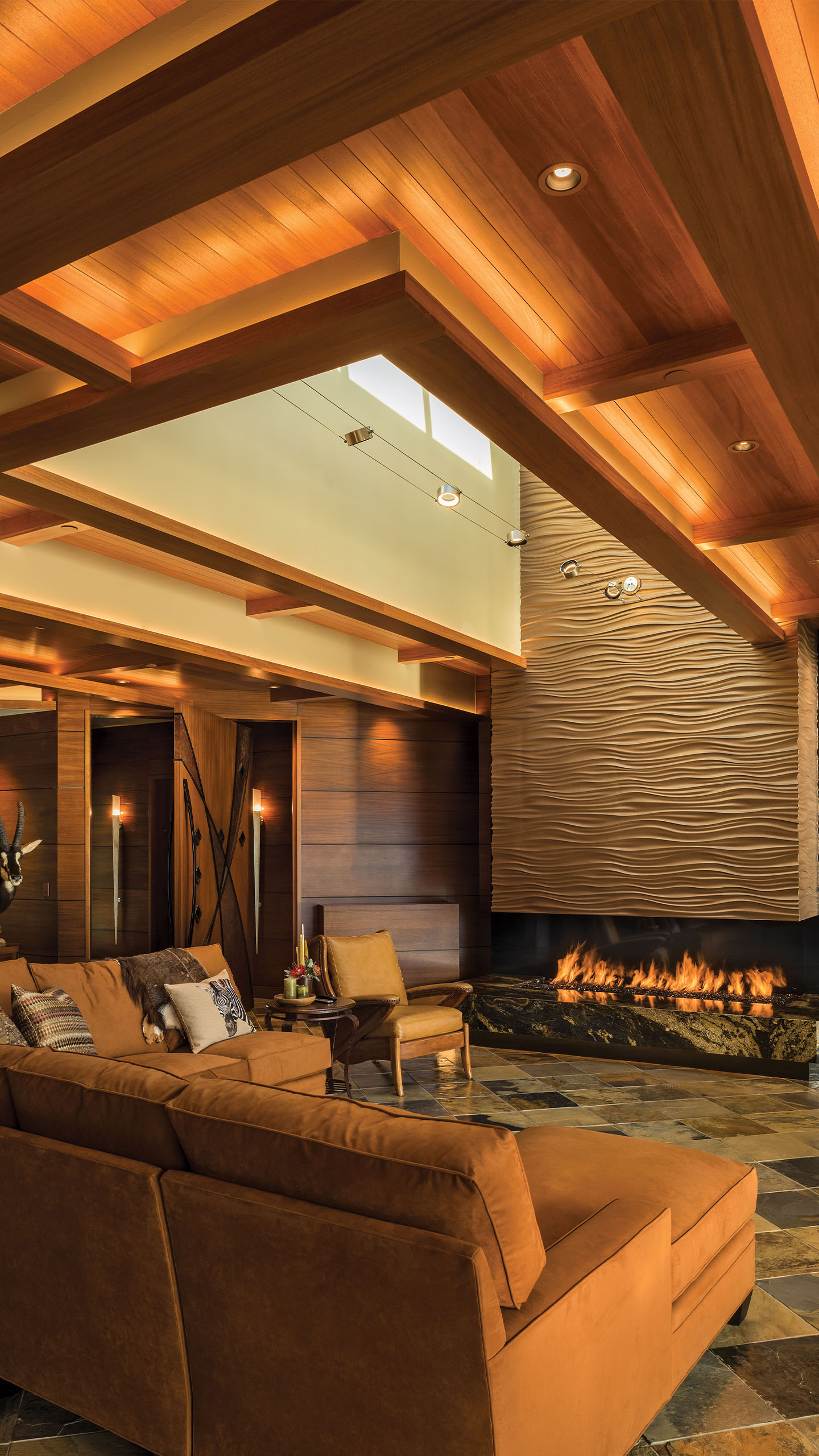 Warm, luxurious living room in the Terrace Condo in Coeur d’Alene, Idaho, featuring a sculptural wave-textured fireplace, rich wood ceiling beams with integrated lighting, and slate flooring. A burnt orange sectional sofa and leather accent chairs create a cozy seating area, while refined materials and layered lighting contribute to a sophisticated atmosphere.