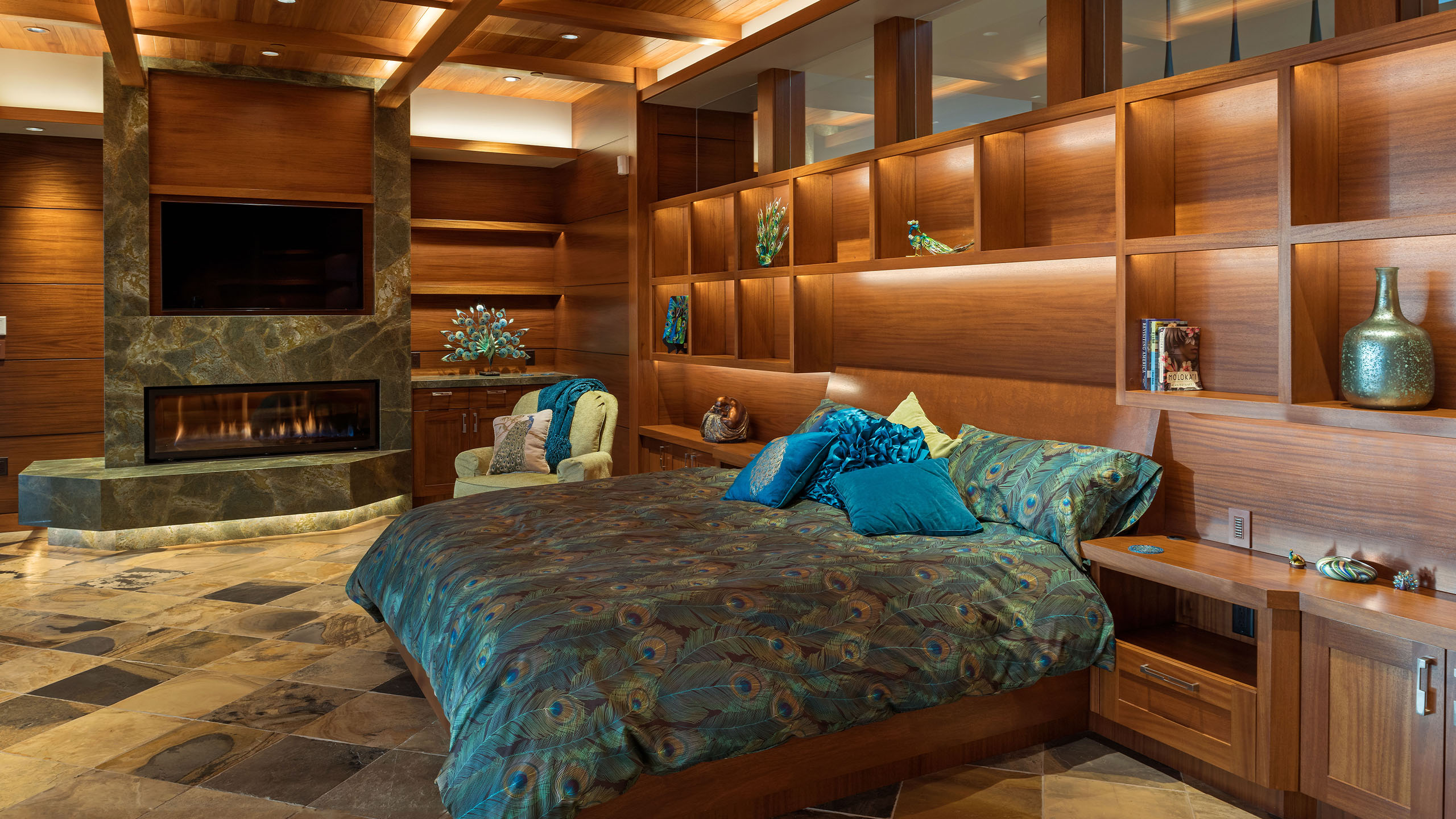 Luxurious bedroom in the Terrace Condo in Coeur d’Alene, Idaho, featuring custom wood cabinetry and shelving, a stone-clad fireplace with built-in TV, and a peacock-inspired bedspread. The space is accentuated by a warm wood coffered ceiling, ambient lighting, and natural stone flooring that enhances the rich, earthy palette.