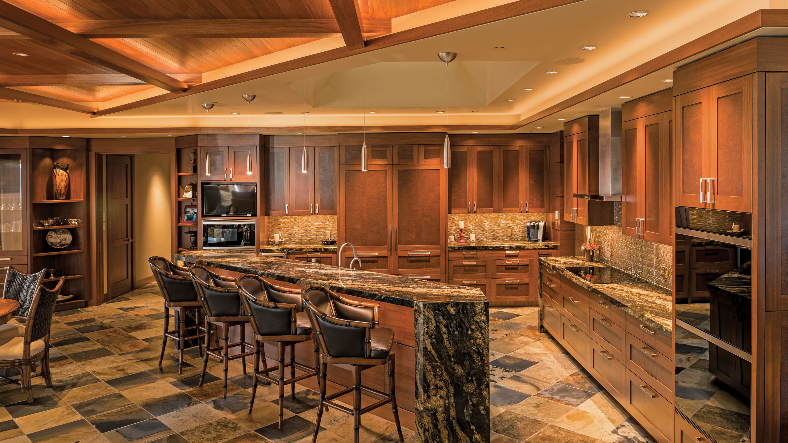 Warm, luxurious kitchen in the Terrace Condo in Coeur d’Alene, Idaho, featuring rich wood cabinetry, a dramatic granite island with bar seating, and slate flooring. A wood coffered ceiling with ambient lighting adds architectural interest, while pendant lights and built-in appliances complete the high-end, elegant design.
