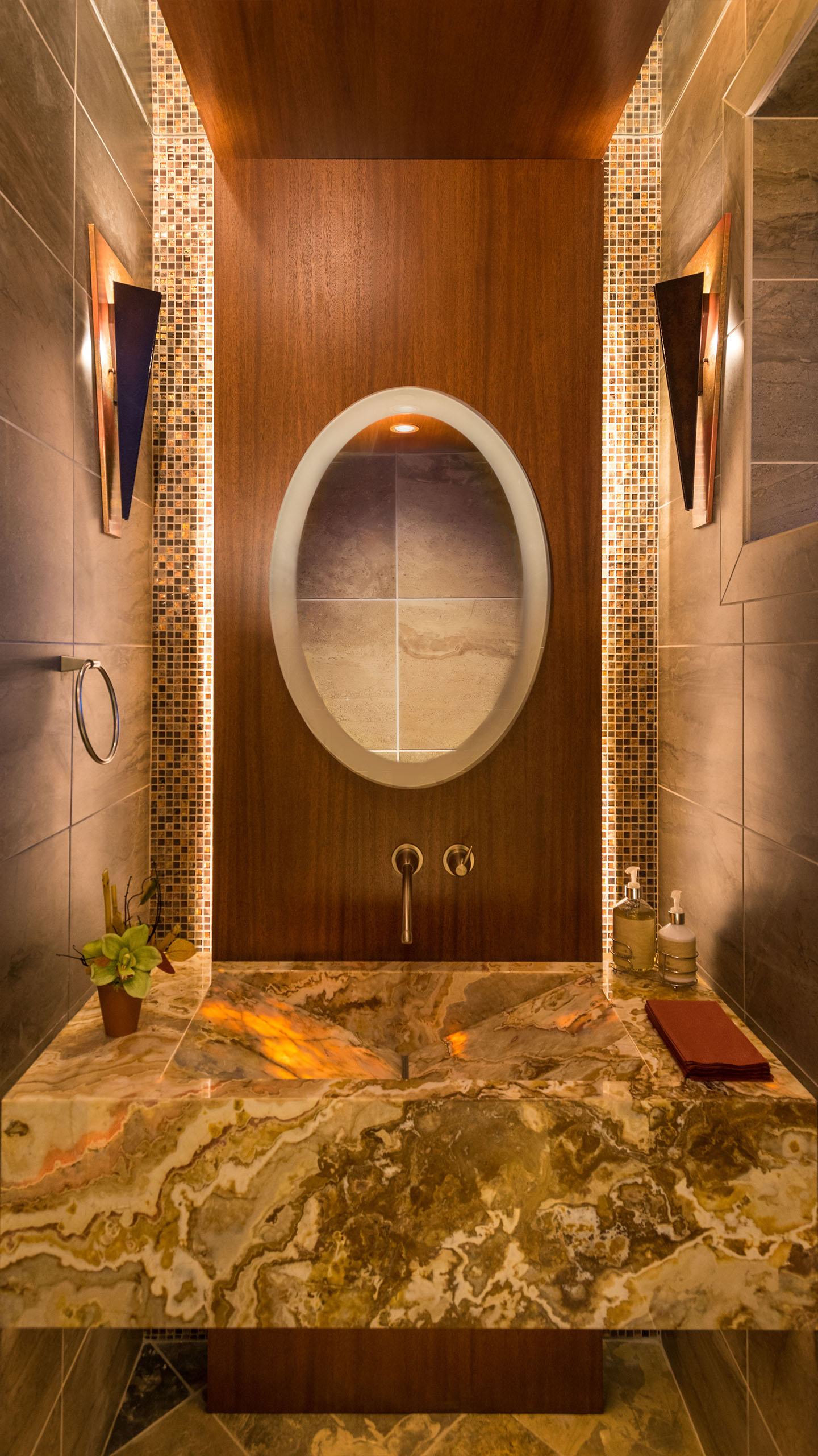 Luxurious powder room in the Terrace Condo in Coeur d’Alene, Idaho, featuring a striking onyx stone sink with warm backlighting, a wood accent wall with an oval mirror, and mosaic tile inlays. Modern wall-mounted fixtures and ambient lighting create a sophisticated, spa-like atmosphere.