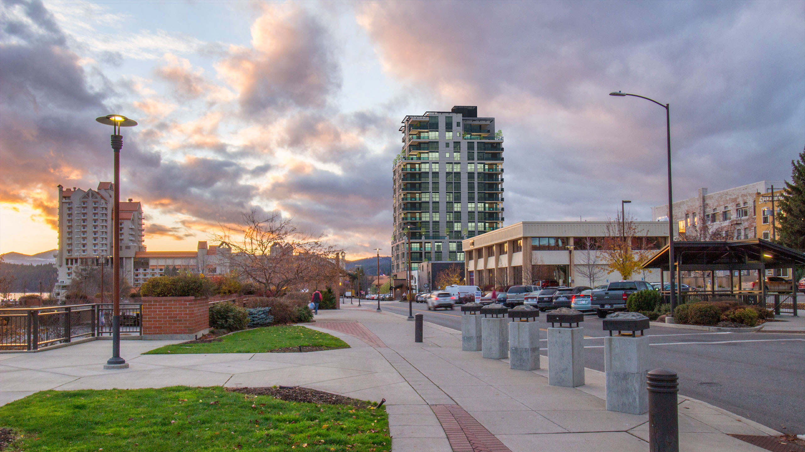 Sunset view of The Thomas George condominium tower in downtown Coeur d’Alene, with pedestrian walkways, nearby buildings, and a dramatic sky.